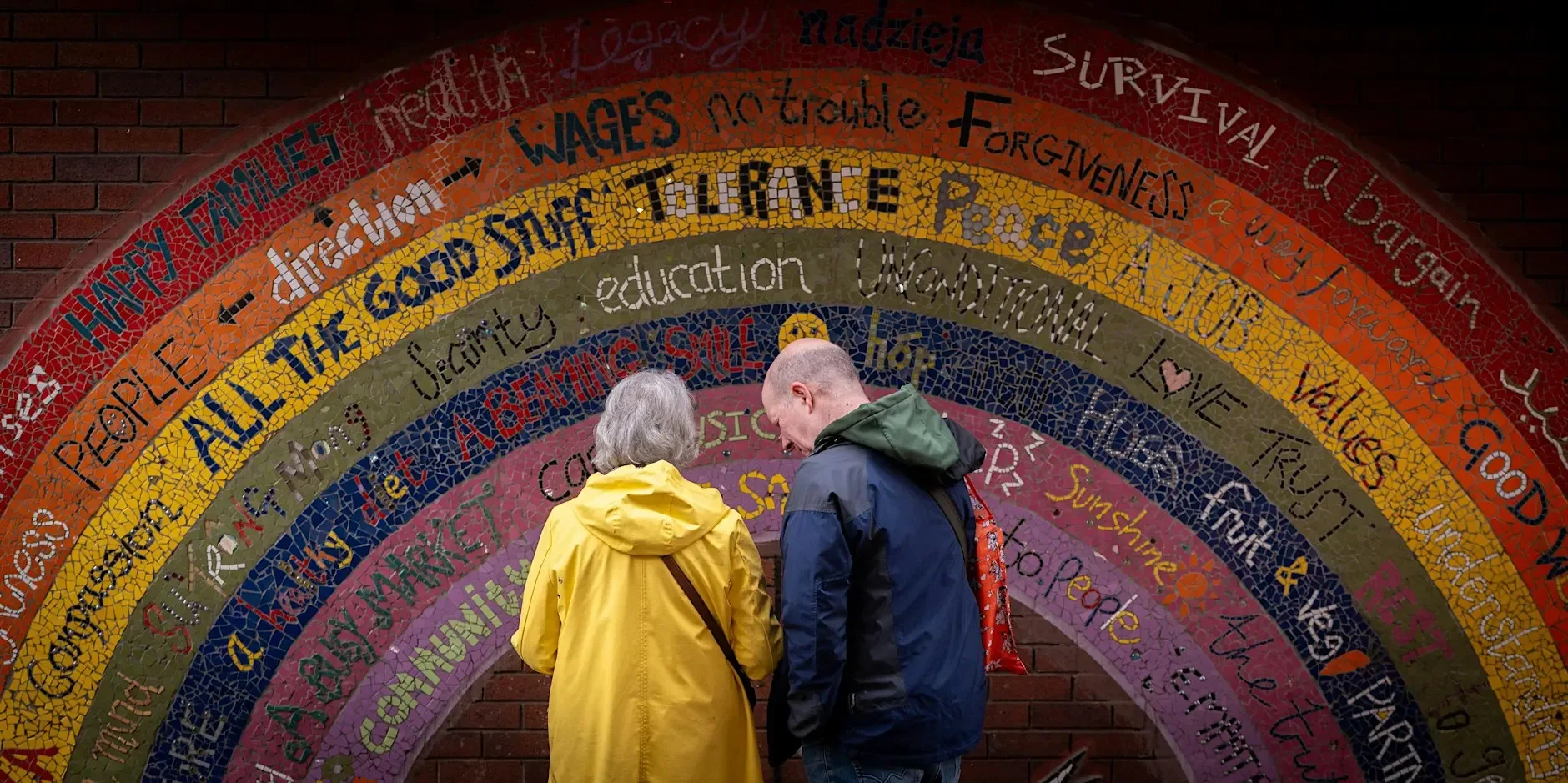 Two visitors standing beneath a colourful mosaic arch covered with words and messages.